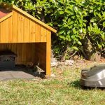 Ein Mähroboter fährt über eine Wiese. Am Rand stehen Hecken und Büsche. Davor steht eine Garage mit integrierter Ladestation für den Mähroboter.