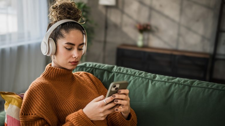 Spotify Playlist uebertragen, Playlists zu Apple Music umziehen Eine Person sitzt im Wohnzimmer auf der Couch, hat Headphones auf und schaut auf ihr Smartphone.