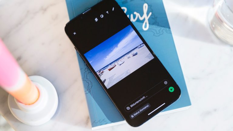 WhatsApp-Status, So funktioniert das Feature Ein iPhone liegt auf einem blauen Notizbuch. WhatsApp ist geöffnet und ein Bild von einem Strand mit Booten soll als neuer Status gepostet werden.