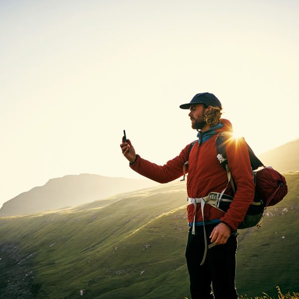 Eine Person steht in Wanderkleidung an einer felsigen Küste und hält ein Smartphone vor sich in der Hand.